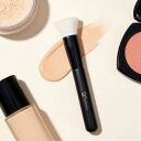 A white, flat-topped Sophienia goat-hair makeup brush, positioned alongside a bottle of liquid foundation, loose powder, and a pink blush compact.