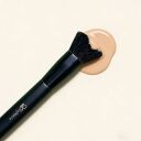 A Sophienia makeup brush, featuring black bristles and a black handle, rests beside a swatch of beige liquid foundation.
