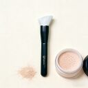 Sophenia makeup brush with white angled bristles and black handle next to loose powder in jar on beige background