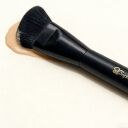 Black angled foundation makeup brush by Sophienia resting on beige liquid foundation swatch against white background