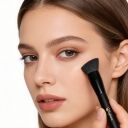 Young woman applying makeup to her cheek with a black Sophienia angled contour brush, natural glowing skin look