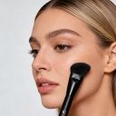 Woman applying makeup with angled contour brush to cheekbone, featuring natural glowing skin and slicked-back blonde hair