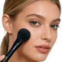 Young woman applying powder to her cheek with a black Kryolan makeup brush, natural makeup look with slicked-back blonde hair