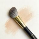 Sophic makeup brush with soft gray bristles and gold ferrule on black handle, resting on peachy powder swatch