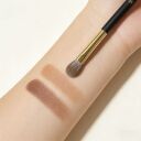 Makeup brush with black handle and gold ferrule resting on forearm showing three brown eyeshadow swatches in warm tones