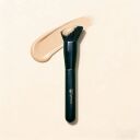 Sophienna makeup brush with dark green handle next to a swatch of beige liquid foundation on cream background
