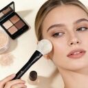 Woman applying loose powder to cheek with fluffy white makeup brush, eyeshadow palette and setting powder in background