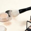 Makeup brush dusting loose powder with cosmetic products including foundation, eyeshadow palette, and setting powder jars