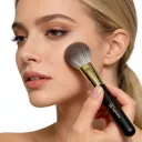 Woman applying blush to cheek with Sophienia makeup brush featuring black handle and gold ferrule