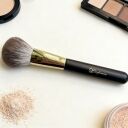 Sophienia makeup powder brush with black handle and gold ferrule surrounded by loose powder and cosmetic palettes