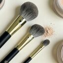 Three makeup brushes with black handles and gold ferrules in varying sizes beside loose powder and compact on cream backgr...