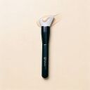 Sophienia foundation brush with flat angled white bristles and black handle, spreading beige liquid makeup on cream background