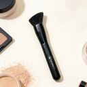 Black angled makeup brush by Sophienia surrounded by loose powder, compact, and cosmetic jars on cream background